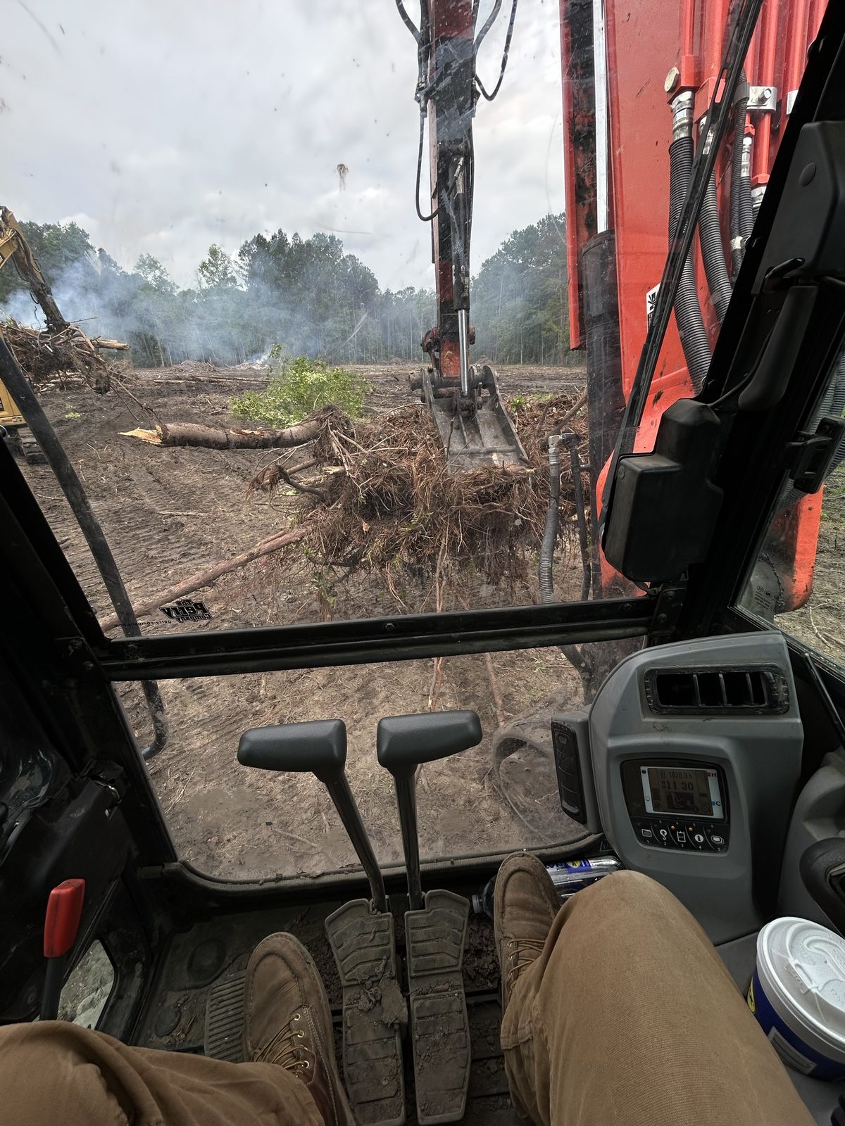 From the excavator cab: the bucket working a pile of roots and brush across a cleared tract, with a second excavator at the tree line