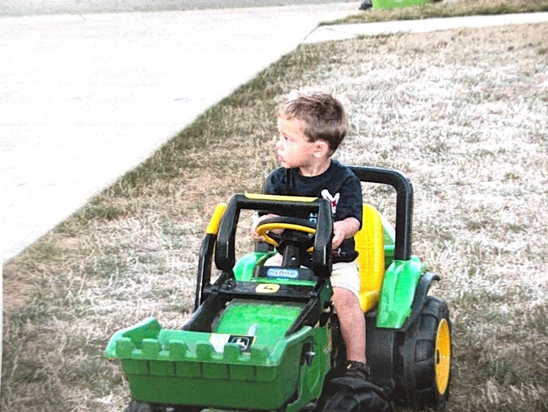 Josiah as a young boy driving a toy tractor in the front yard